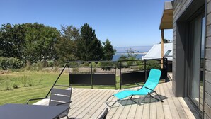 Eine Holzterrasse mit Tisch und Stühlen, ein blauer Liegestuhl und Meerblick.