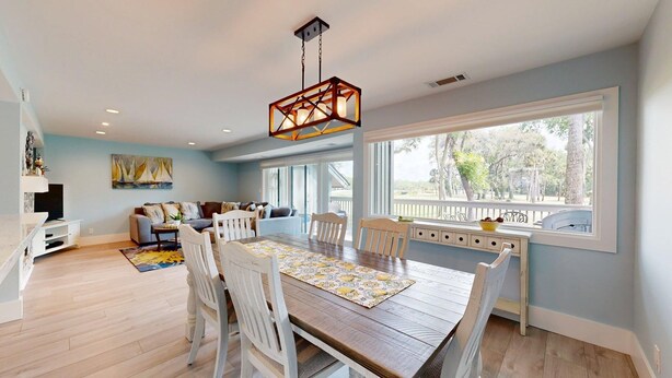 A spacious living room with a large window, a dining area with a wooden table and white chairs, and a ceiling light fixture. - Large Three Bedroom Villa Directly on George Fazio Championship Golf Course! (Hilton Head)