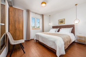 Ein Schlafzimmer mit einem Bett, einem Sessel, einem Holzschrank, einem Fenster mit Vorhängen und einer Hängeleuchte.