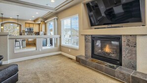 Amplia sala de estar con chimenea, un televisor grande montado encima y una cocina visible a través de un arco abierto.