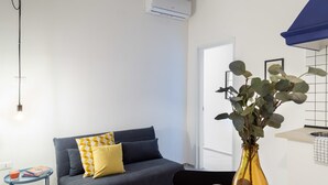 Uma sala de estar moderna com sofá, mesa lateral pequena, um vaso com plantas e uma área de cozinha com pia e bancada.