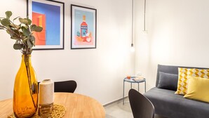 Uma sala de estar moderna com uma mesa redonda de madeira, um sofá cinza com almofadas estampadas em amarelo e branco, e duas obras de arte emolduradas na parede.