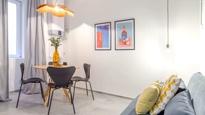 Uma sala de estar moderna com um sofá cinzento, uma mesa de jantar com cadeiras e uma luminária pendente. Há duas molduras com quadros na parede e um vaso com uma planta na mesa.