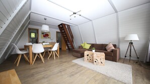 A modern loft-style living space with a wooden floor, a brown sofa, white chairs, a dining table, and a staircase.
