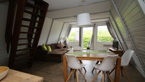 A cozy, well-lit attic bedroom with a wooden ladder, a bed, a desk, and a dining area with chairs and a table.
