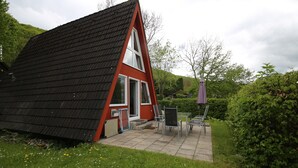 A small, red, A-frame house with a black roof, a patio with chairs, and a garden.