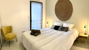 A neatly arranged bedroom with a large bed, a chair, a wall-mounted woven artwork, and bedside lamps. - Appartement T3 de Standing - 2 Chambres 4/6 Personnes Porto Vecchio (Porto-Vecchio)