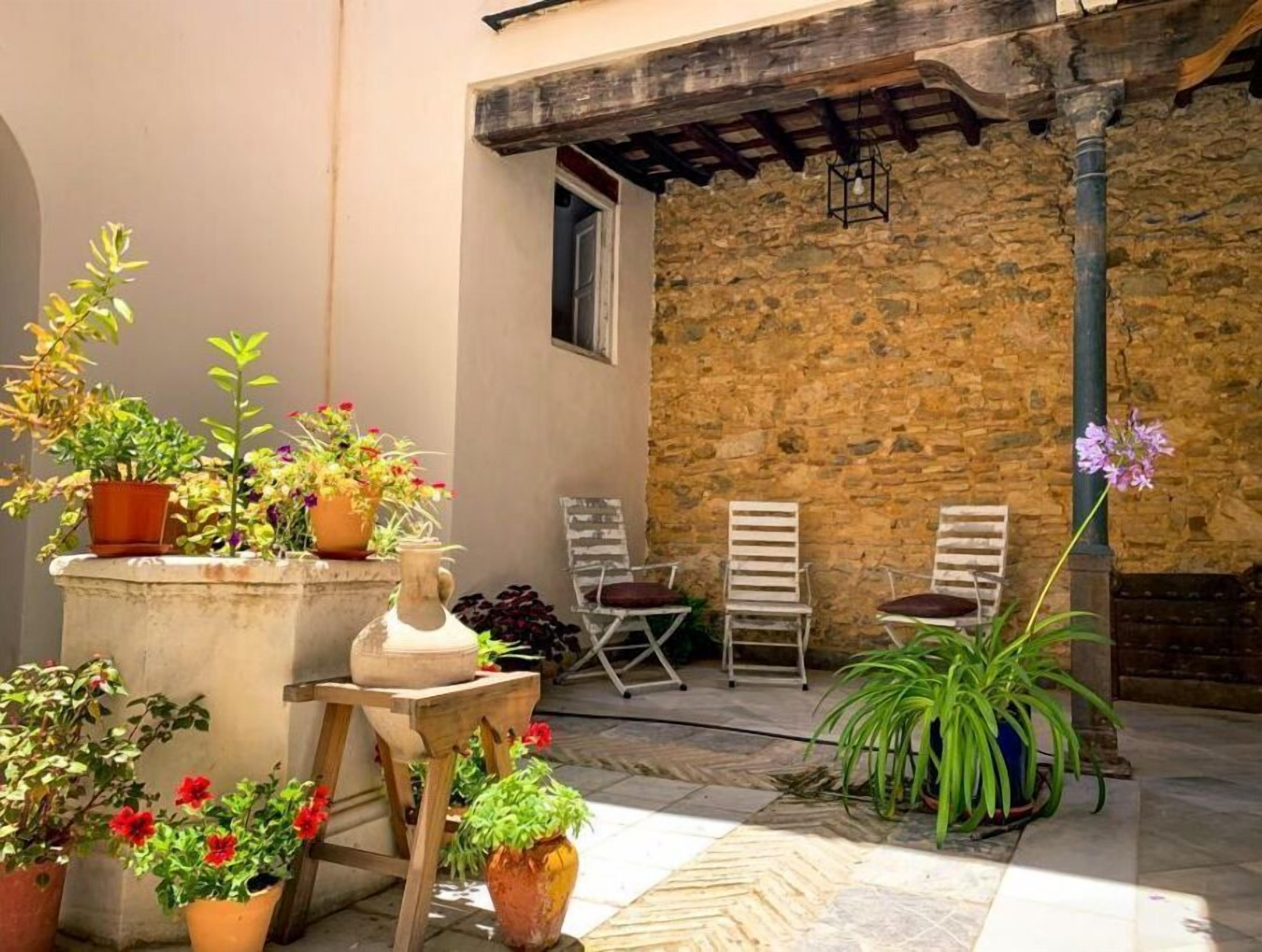 A courtyard with potted plants, two white chairs, and a stone wall.
