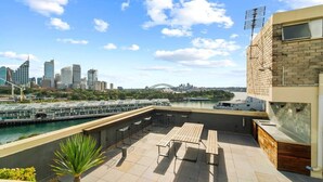A rooftop terrace with a view of the Sydney Opera House and Harbour Bridge, featuring outdoor seating and a modern building facade.