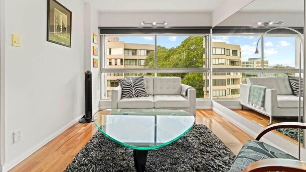 A modern living room with a glass coffee table, grey sofas, and a large window offering a city view.