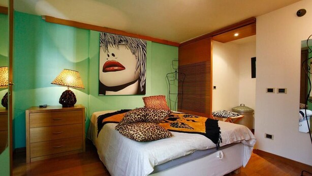 A bedroom with a bed, a nightstand, a lamp, a wall art piece, and a window with blinds. - Casa Vacanza Diana (roma)