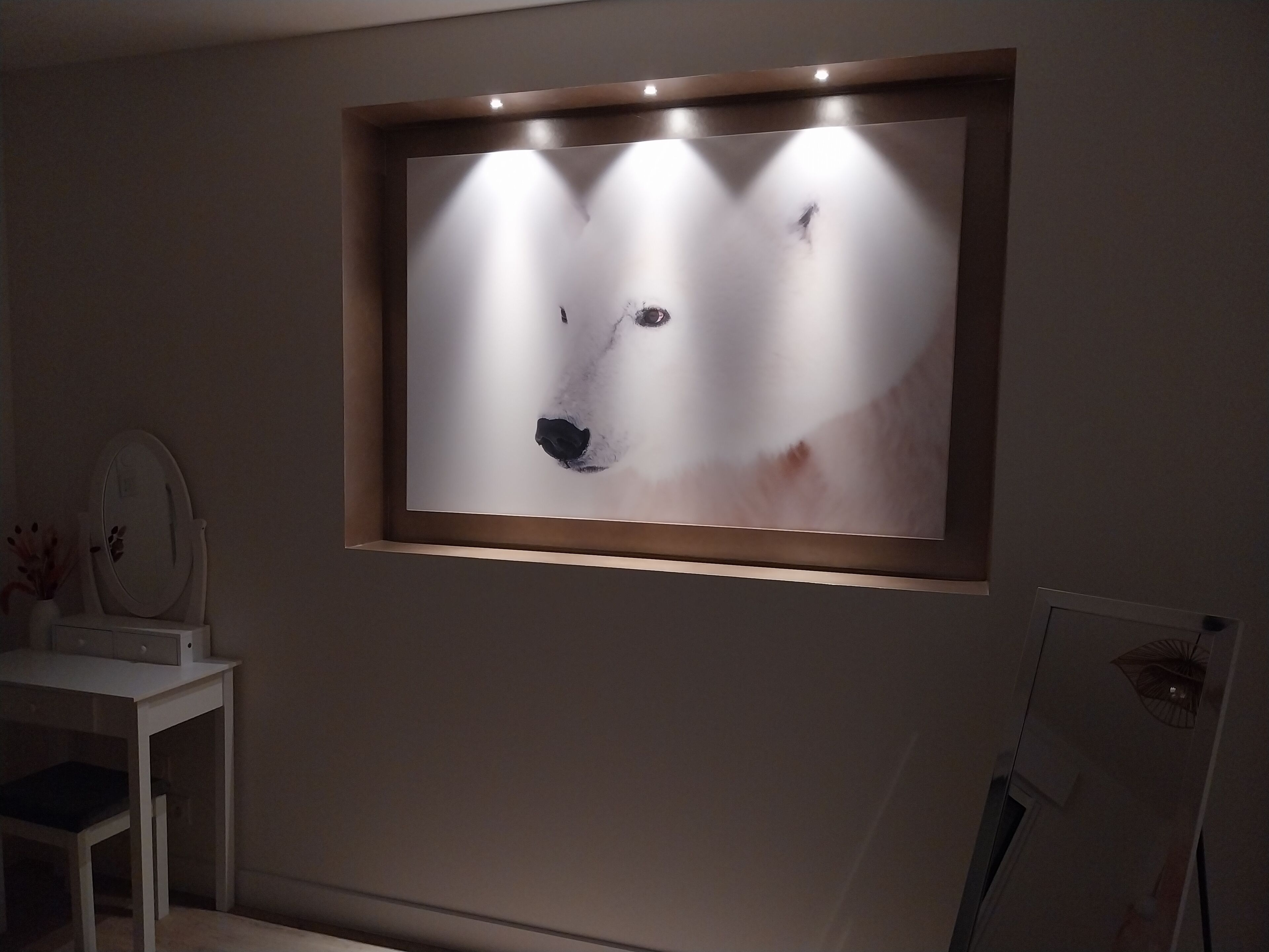Una obra de arte en un marco sobre una pared, una mesita con un espejo y una silla.
