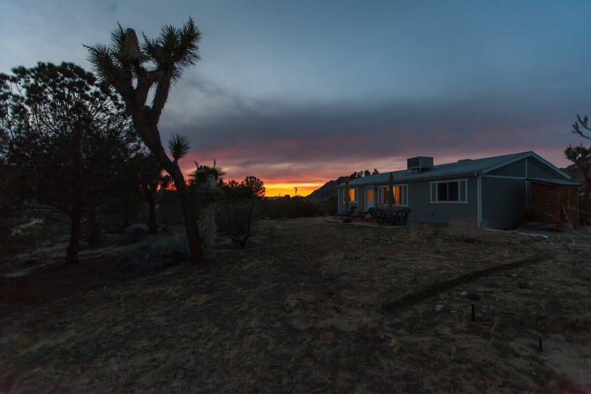 A modern house with a large yard, surrounded by trees and a sunset in the background.