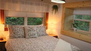 A bedroom with a bed, two bedside lamps, a window with a view of greenery, and a hanging plant.