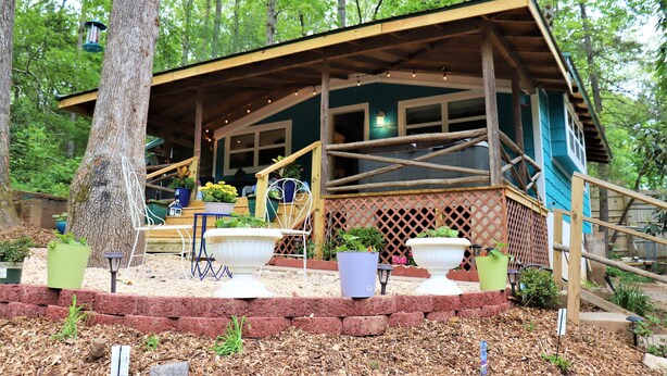 A rustic cabin with a wooden deck, surrounded by trees and potted plants.