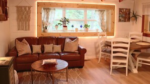 A cozy living room with a brown leather sofa, a round coffee table, and a dining table with white chairs. There is a window with plants on the sill and a wall-mounted macrame piece.