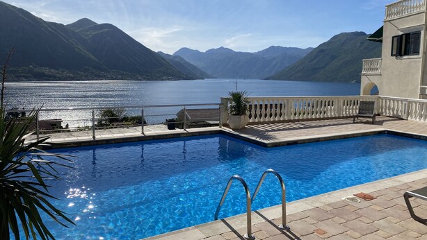 En swimmingpool med kristallklart blått vatten, omgiven av en kakelbeklädd terrass och ett vitt räcke. I bakgrunden syns en sjö och berg.