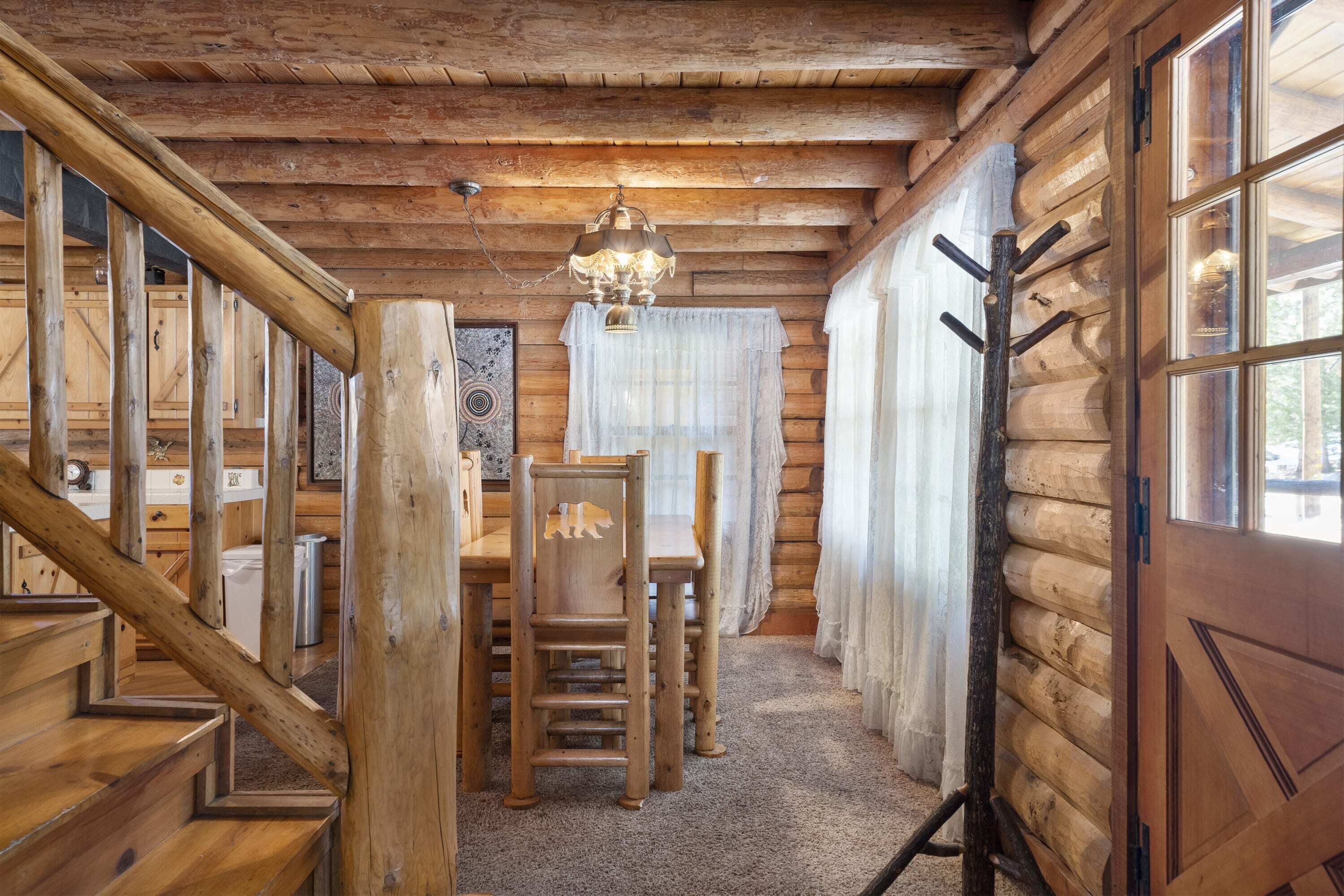 Interno di una baita in legno con una scala in legno, un tavolo da pranzo con sedie, un lampadario e un'ampia finestra con tende.