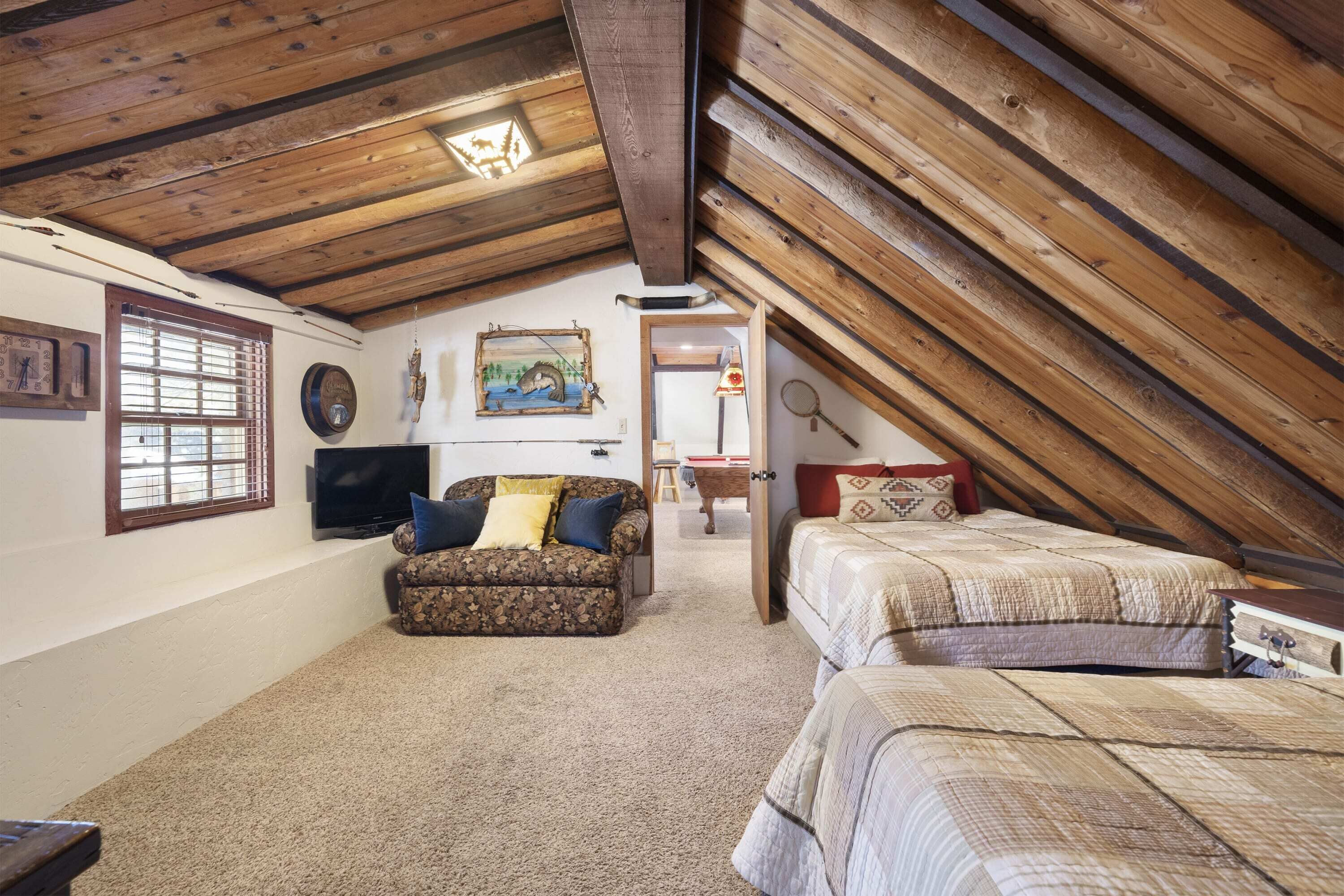 A cozy attic bedroom with a wooden ceiling, a sofa, two beds, a television, and a painting on the wall.