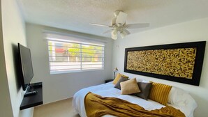 A bedroom with a bed, a ceiling fan, a TV mounted on the wall, a window with blinds, and a wall art piece.