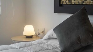 Una cama bien hecha con un almohadón gris y una lámpara de mesa encendida a su lado.