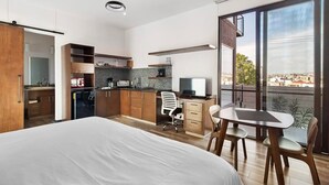 Une chambre d’hôtel moderne dotée d’un grand lit, d’un bureau avec une chaise, d’une kitchenette et d’un balcon offrant une vue sur la ville.