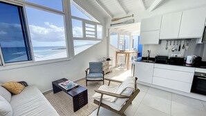 A modern living room with a sofa, armchair, and coffee table, a kitchen with white cabinets and a dining area, and large windows with a view of the ocean. - Deep Blue Vue mer et Piscine Privée (Le Gosier)