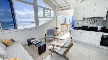 A modern living room with a sofa, armchair, and coffee table, a kitchen with white cabinets and a dining area, and large windows with a view of the ocean.