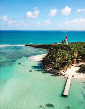 A coastal scene with a lighthouse, clear turquoise waters, a sandy beach, and a pier.