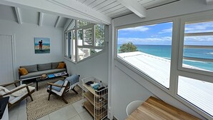A room with a large window overlooking the beach, a sofa, a small table, and a cart with items. - Deep Blue Vue mer et Piscine Privée (Le Gosier)