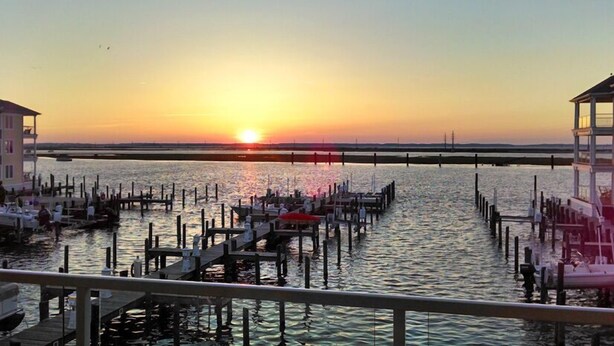 A waterfront scene with a pier extending into calm water, a sunset in the background, and a bridge in the distance. - Bayside & Beyond- 1st Floor Waterfront Condo with Pool, Gym & Boat Slip (Chincoteague)