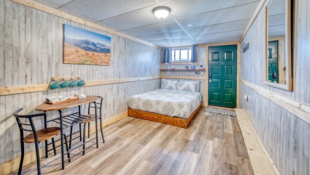 A bedroom with a wooden floor, a bed with a white bedspread, a desk with chairs, a painting on the wall, and a green door.