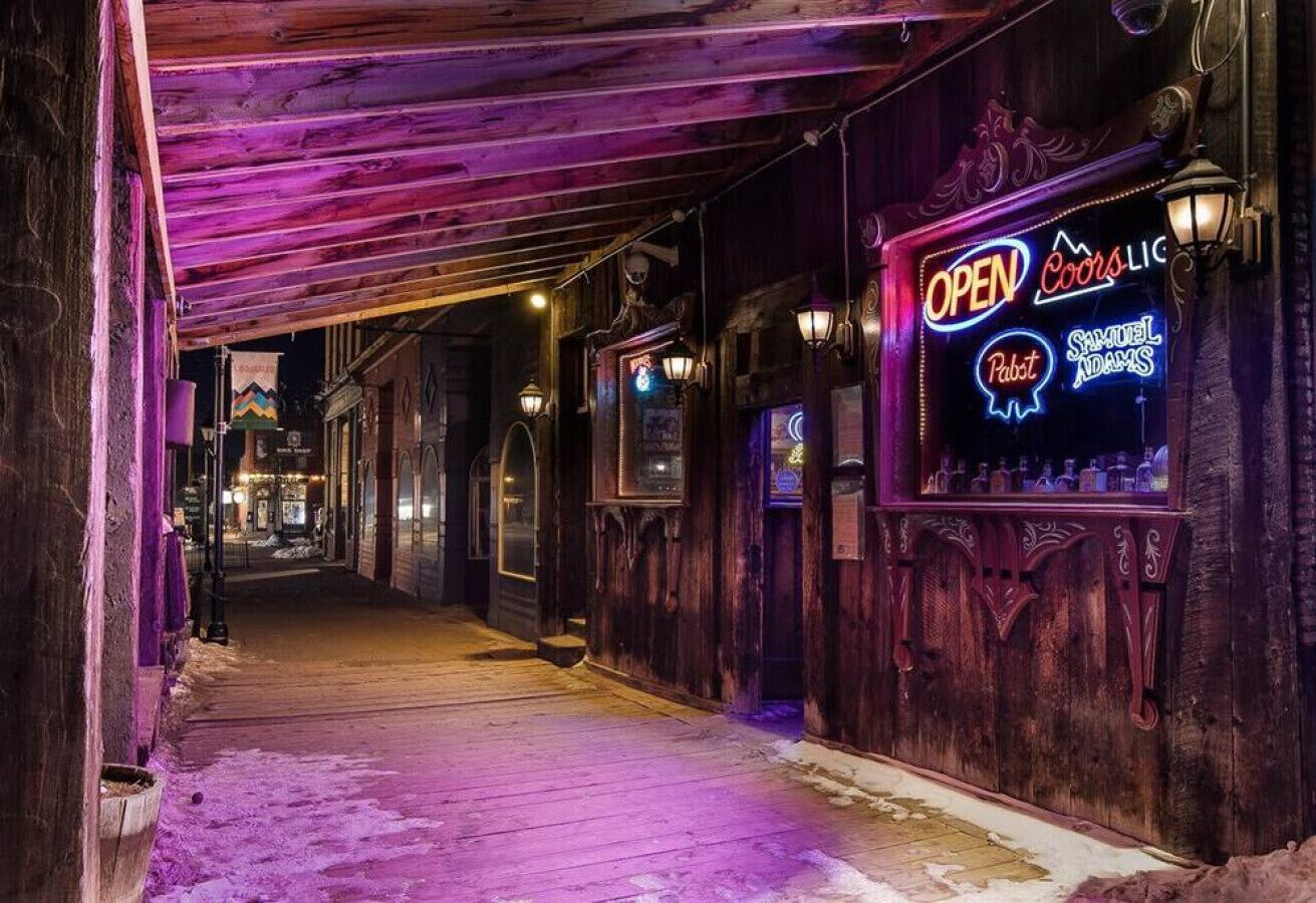 Ein schmaler Korridor mit Holzwänden und einem leuchtenden Neon-Schild, das eine Bar namens „OPEN Coors“ anzeigt.