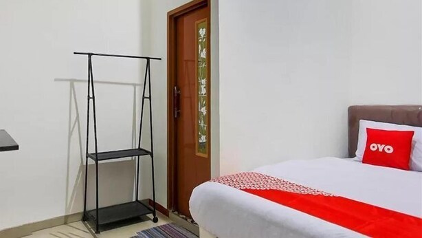 A bedroom with a bed, a red pillow, a white bedspread, a black metal shelf, and a wooden door with green decorations. - OYO 3456 Citara Jaya Guesthouse (Bekasi)