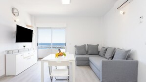 Una sala de estar moderna y luminosa con una mesa de comedor blanca, sofá gris y televisor montado en la pared.