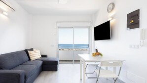 Ein modernes Wohnzimmer mit grauem Sofa, einem weißen Esstisch mit Stühlen, einem Flachbildfernseher, einer Uhr und Blick auf den Strand durch ein großes Fenster.