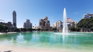 Ein Strand mit einer Fontäne, klarem Wasser und Gebäuden im Hintergrund.
