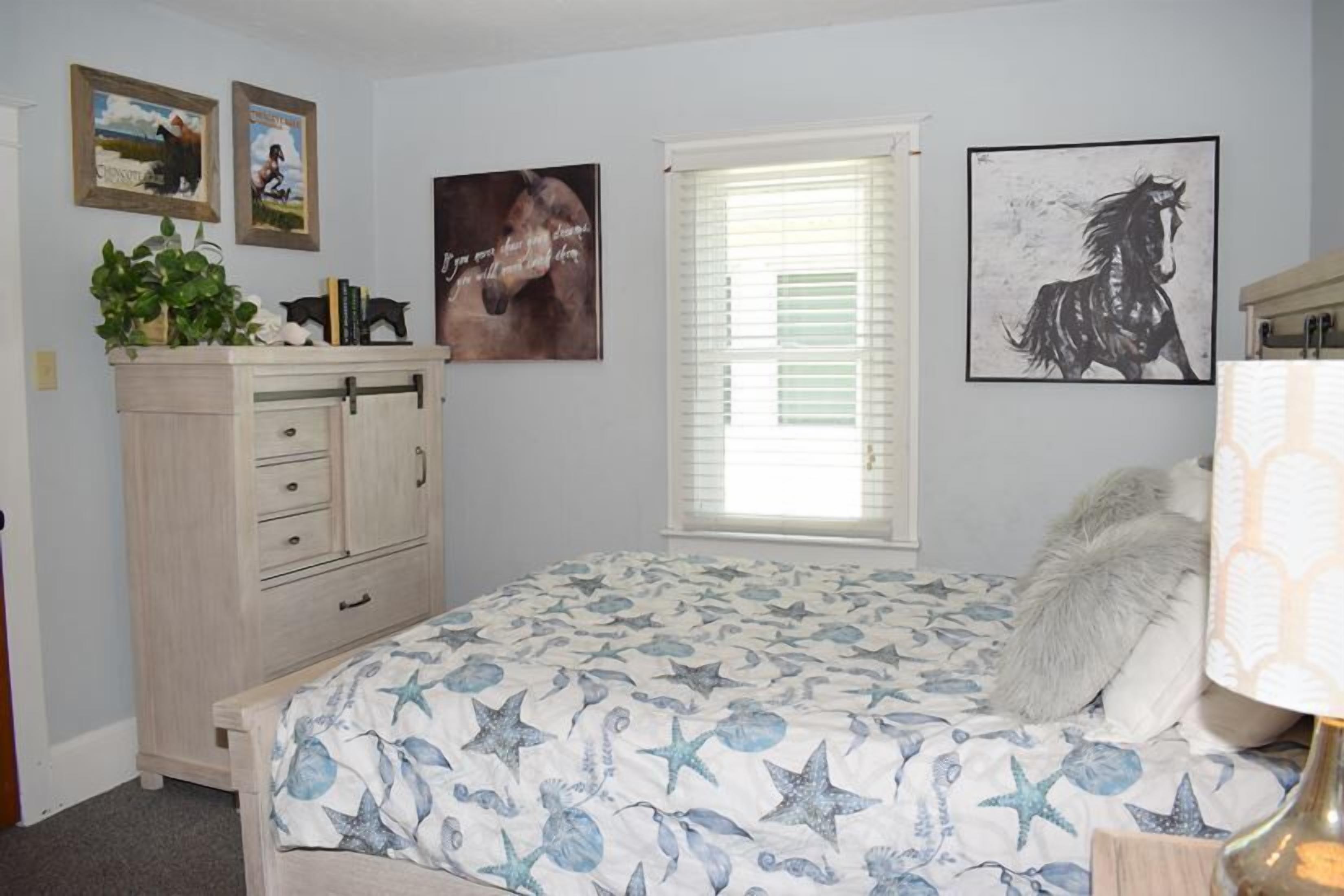 Une chambre à coucher comprenant un lit, une commode, une fenêtre avec des stores et des œuvres d’art murales.