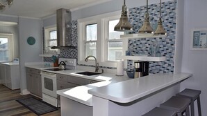 Eine moderne Küche mit weißen Arbeitsplatten, einer Edelstahl-Dunstabzugshaube und einer blau gefliesten Spritzschutzwand. Zwei Pendelleuchten leuchten über der Kochinsel, und ein Fenster lässt natürliches Licht herein.