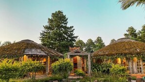 Ein friedlicher Garten mit zwei strohgedeckten Gebäuden, einem zentralen Pavillon und einem Teich.