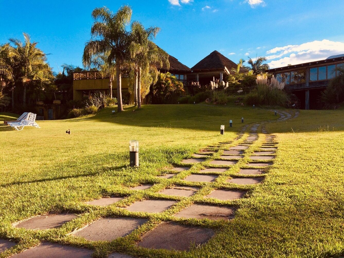 A pathway leading to a building with a thatched roof, surrounded by lush greenery and palm trees.