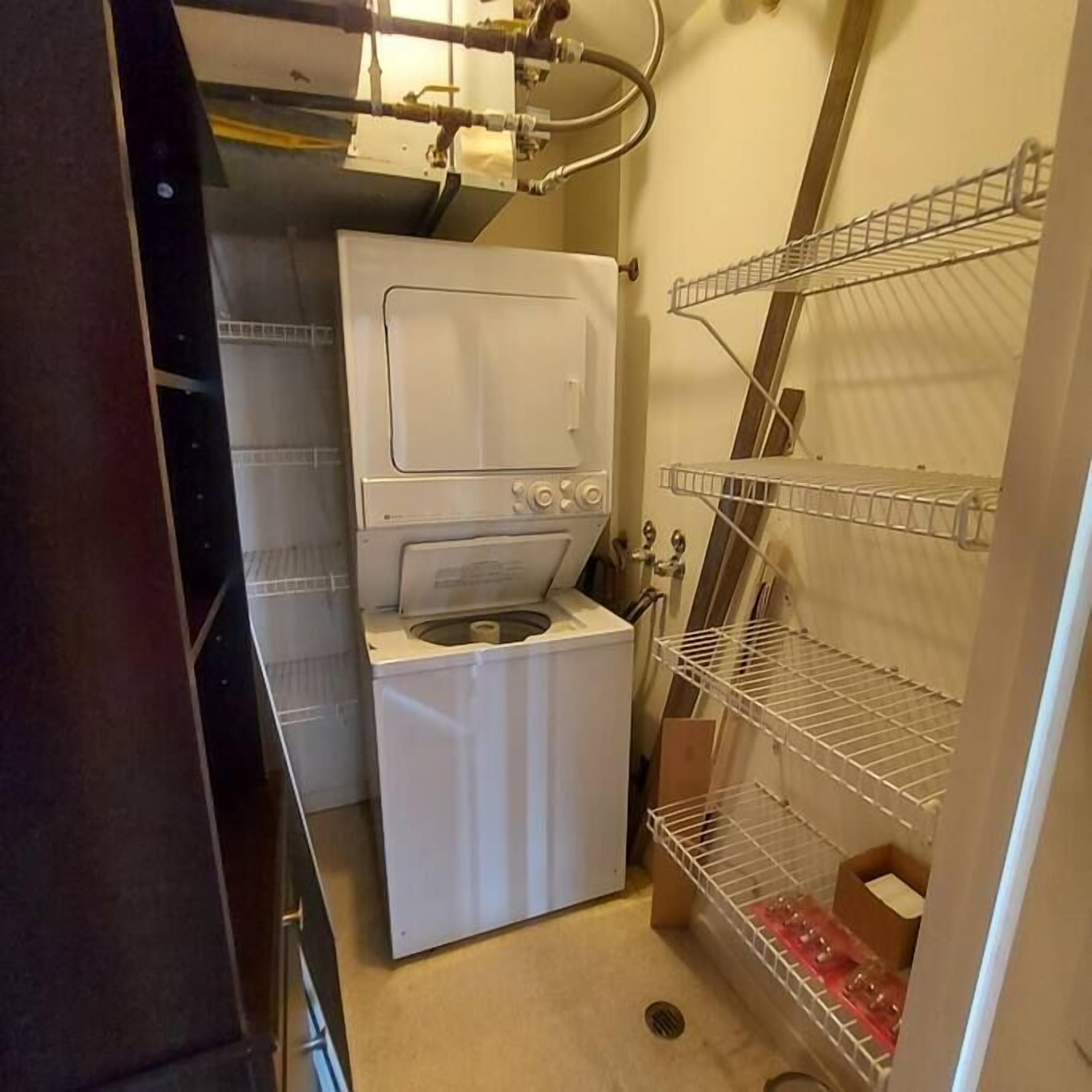 A compact laundry room with a front-loading washer and dryer, metal shelving units, and a visible plumbing system.