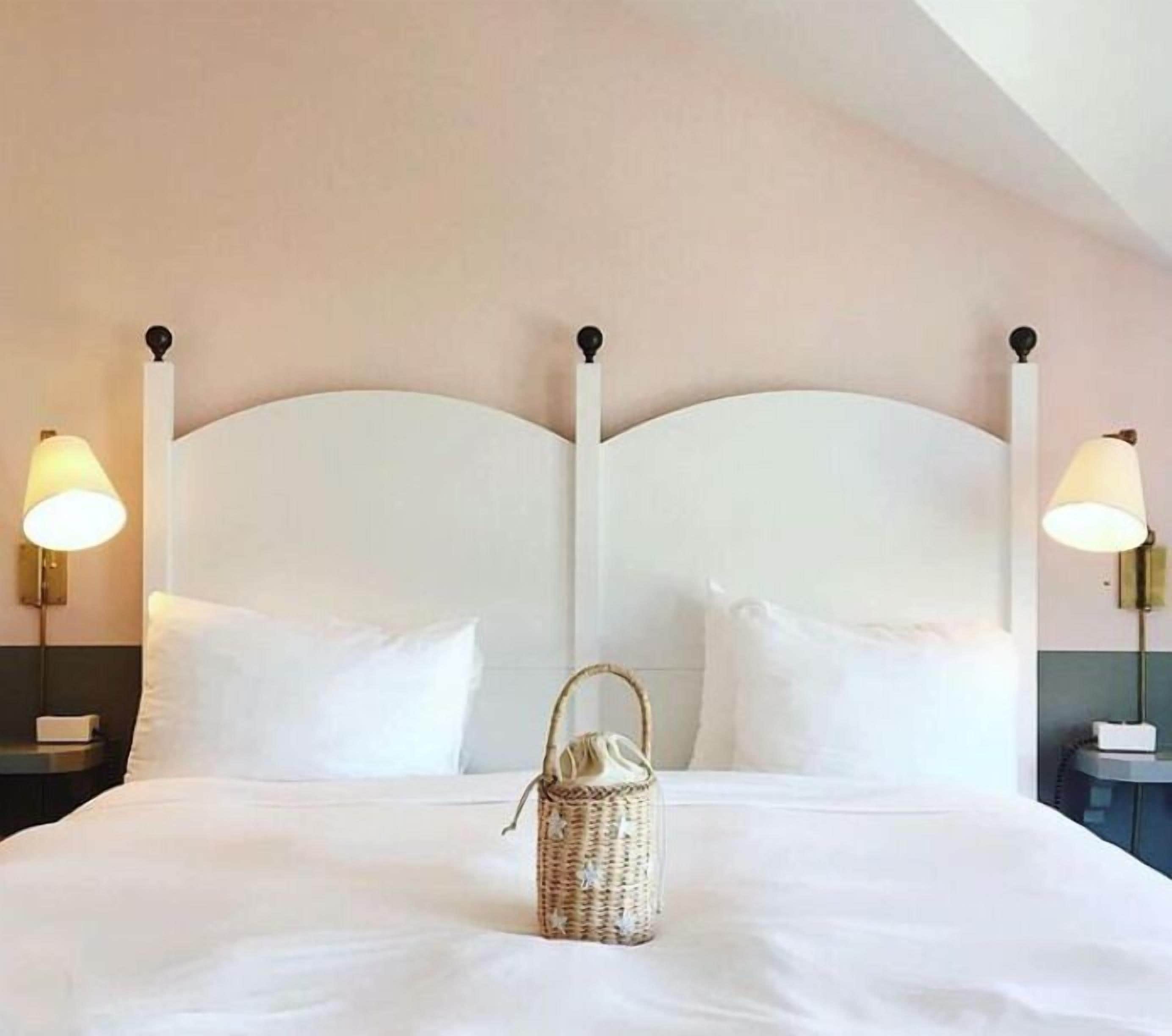Una cama bien hecha con sábanas blancas, una cesta de mimbre sobre la cama y dos lámparas de noche a cada lado.