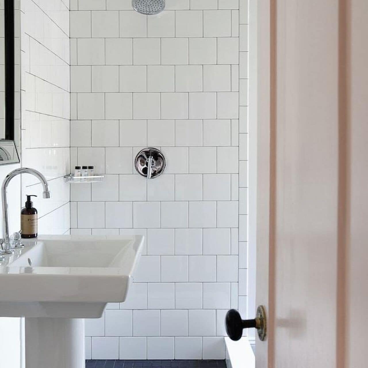 A bathroom with a white tiled wall, a sink, a showerhead, and a door.