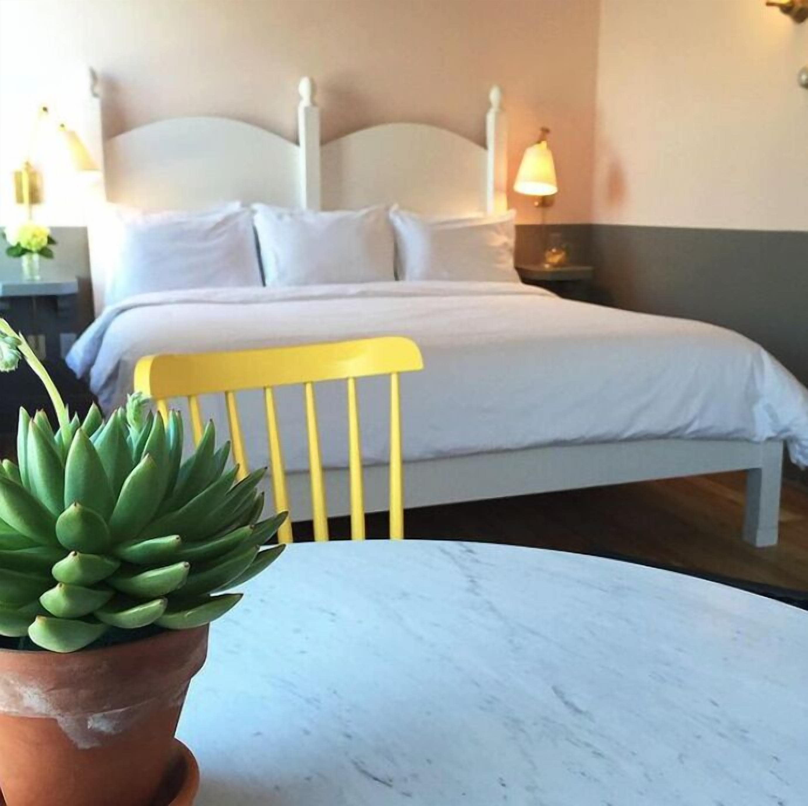 Un lit bien fait, avec du linge de lit blanc, une chaise jaune et une plante en pot posée sur une table en marbre.