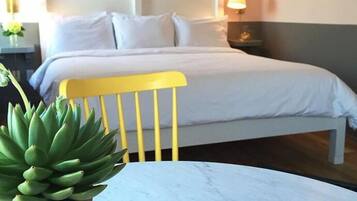 Uma cama bem arrumada com lençóis brancos, uma cadeira amarela e uma planta em vaso sobre uma mesa de mármore.