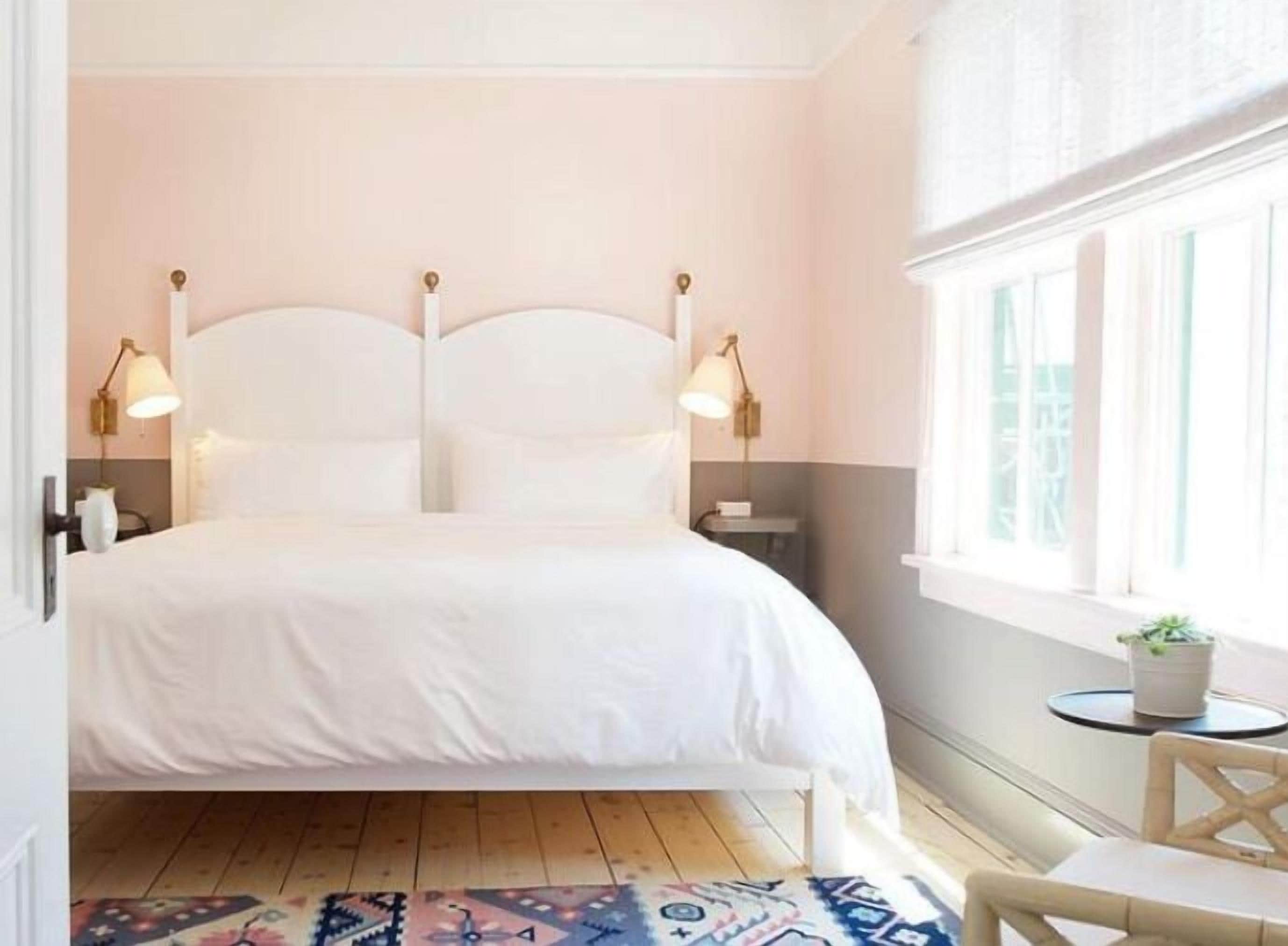 A neatly arranged bedroom with a double bed, bedside lamps, a small table with a plant, and a window with curtains.