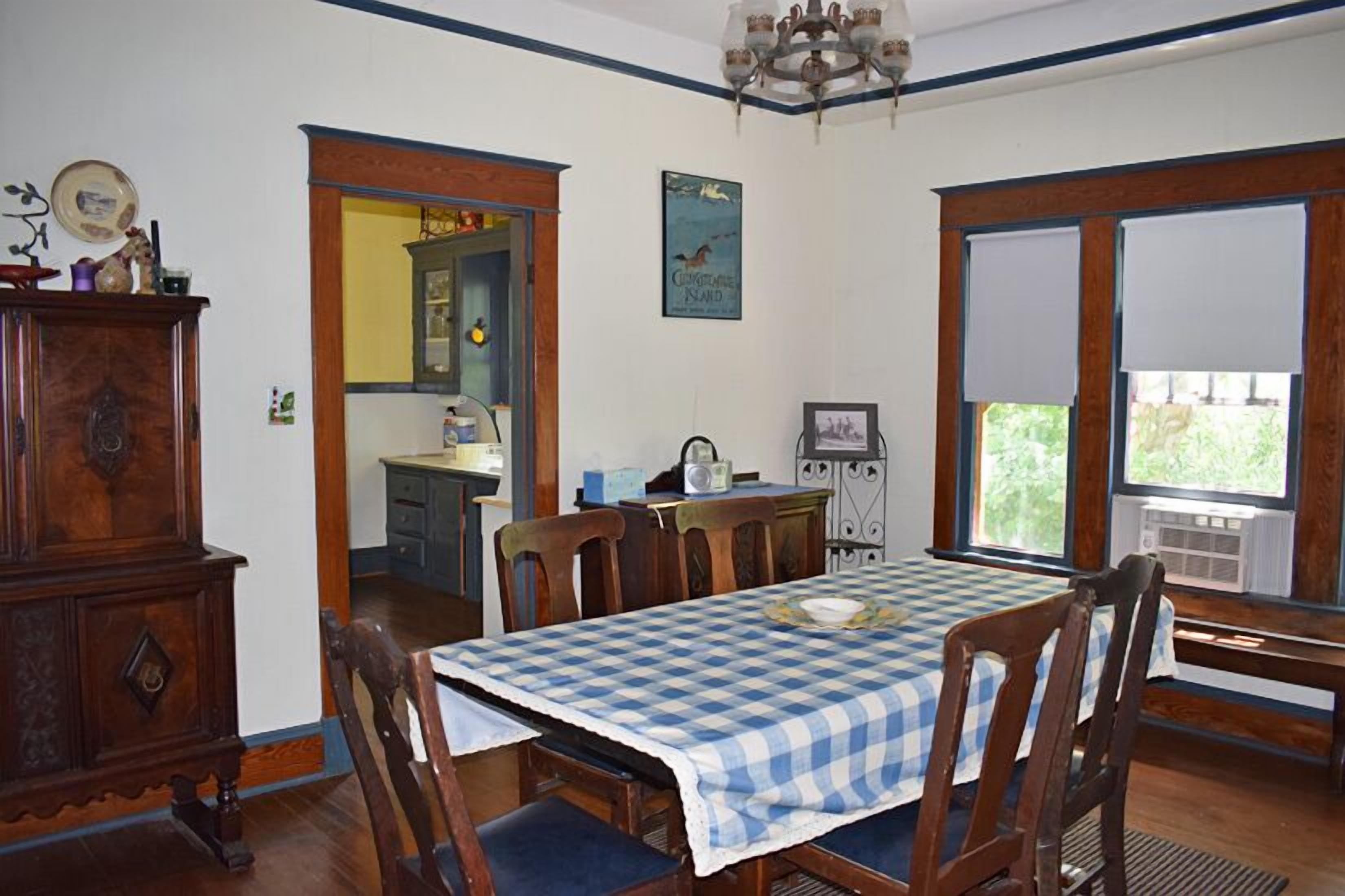 A dining area with a checkered tablecloth, wooden chairs, a chandelier, and a view through large windows.