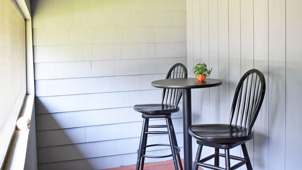 Un pequeño espacio al aire libre con una mesa redonda y dos sillas, una planta en maceta y una pared de madera blanca.