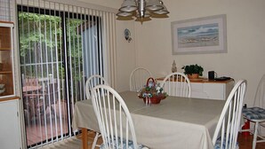 Un comedor con una mesa dispuesta para cuatro personas, rodeada de sillas blancas. Un candelabro cuelga sobre la mesa, y hay un cuadro con pájaros en la pared.